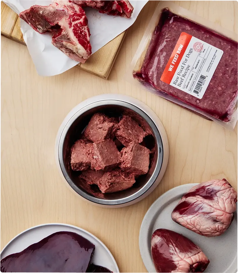 Various cuts of raw meat and prepared raw dog food displayed on a wooden cutting board with butcher knife and kitchen towels. Includes raw muscle meat, organs, and ground meat in a metal container.